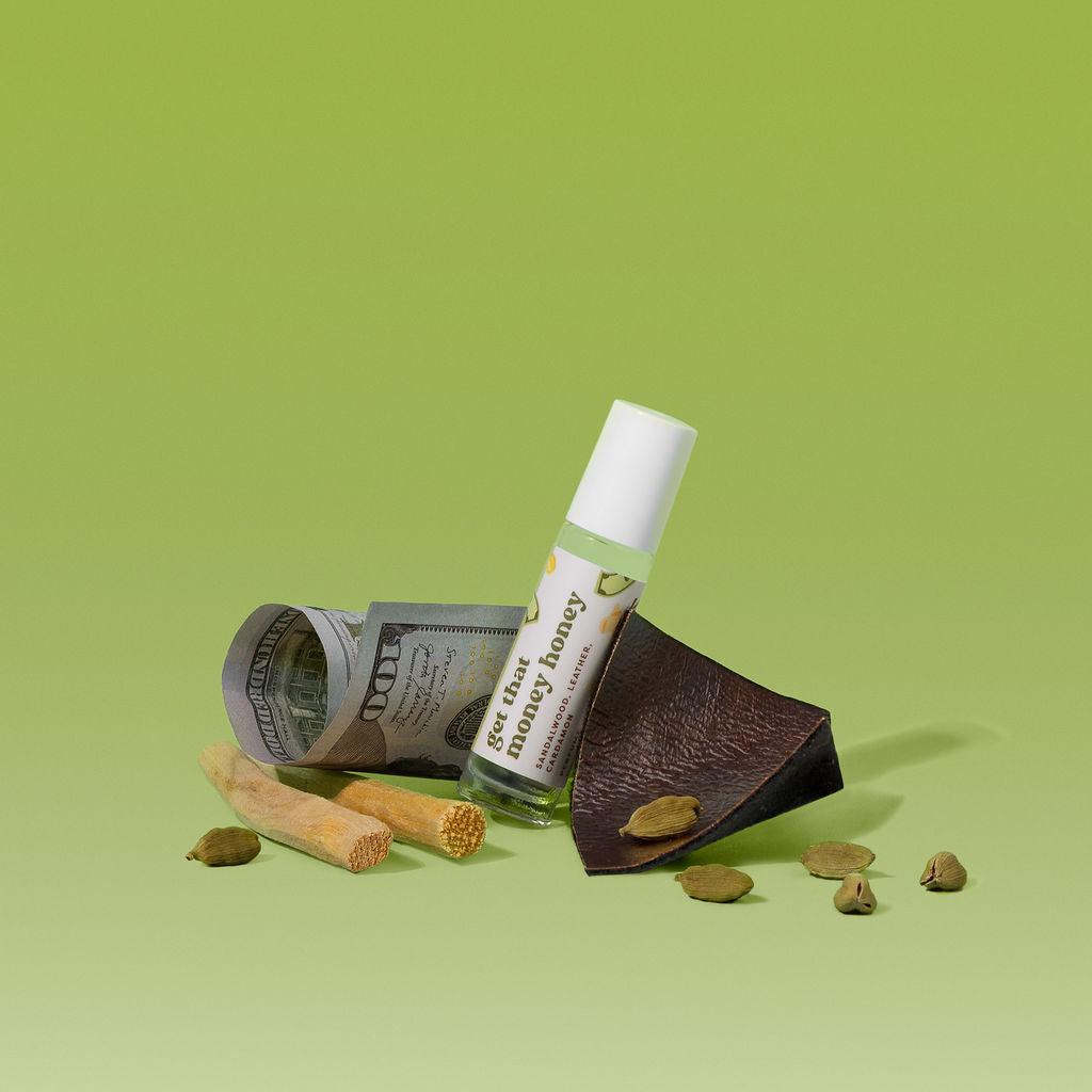 A small clear roll-on bottle labeled 'get that money honey' stands upright beside a rolled dollar bill, a dark brown leather wedge, and several green cardamom pods on a light green background.