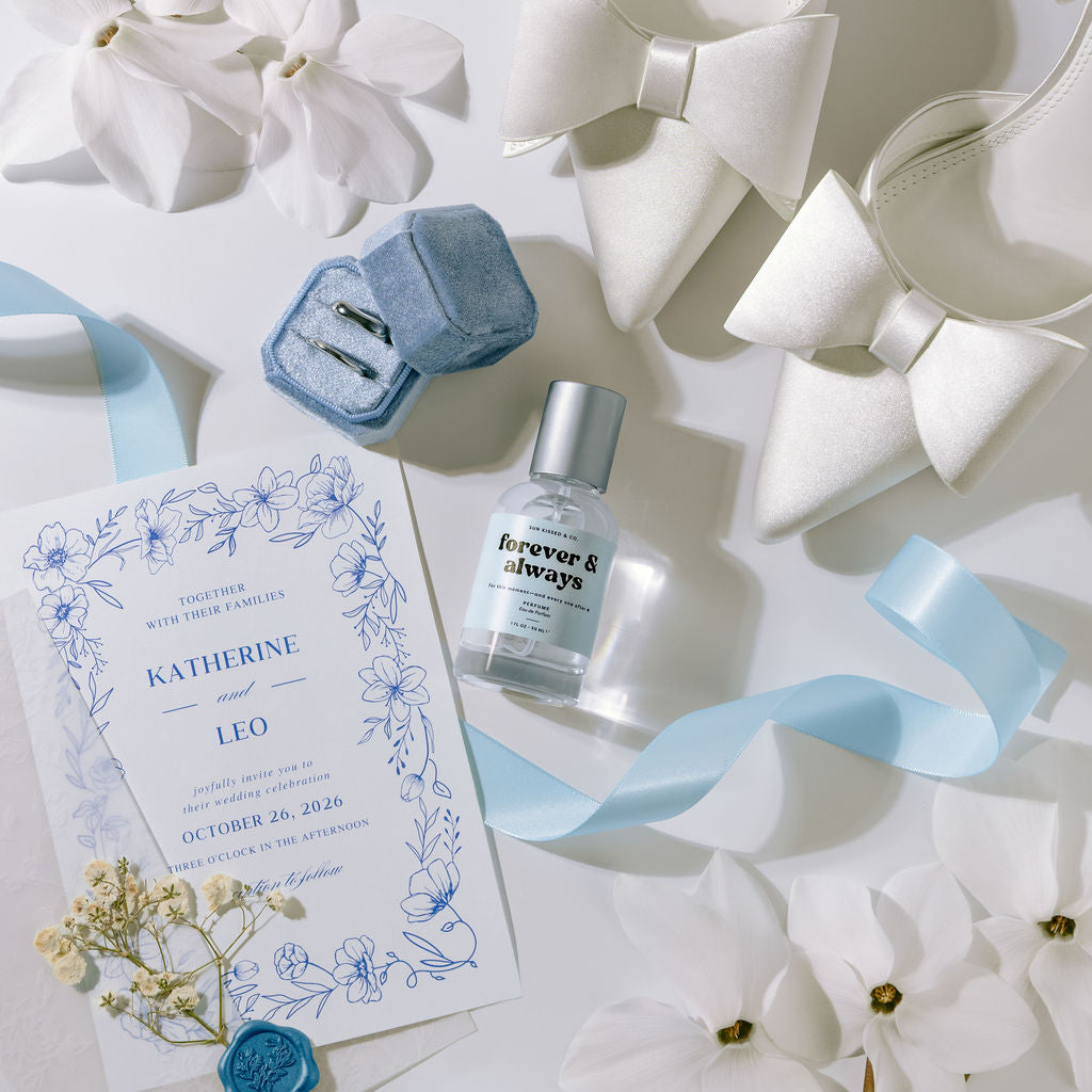 A styled flat-lay featuring a wedding invitation card with blue floral border details, a small bottle of 'forever & always' perfume, a blue velvet ring box containing two rings, and white satin bow headbands, all arranged with white flowers and a light blue ribbon on a white background.