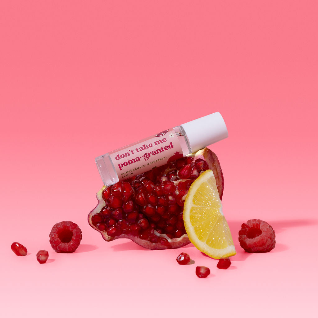A small clear bottle with a white cap labeled 'don't take me poma-granted' rests on top of a pomegranate half, styled with a lemon slice, raspberries, and scattered pomegranate seeds on a pink background.