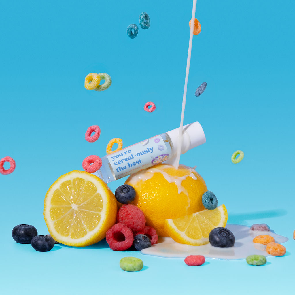 A small clear bottle with a white cap and a colorful label reading 'you're cereal-ously the best' is shown on a bright blue studio background, surrounded by floating cereal pieces, a lemon half, a whole lemon, blueberries, raspberries, and a milk splash. 