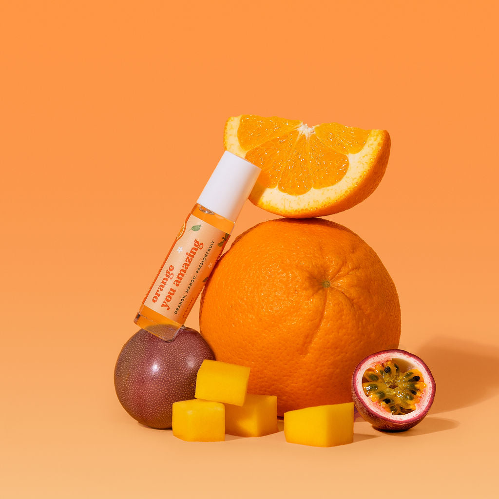 A small roll-on bottle labeled 'orange you amazing stands on a passionfruit, styled with fresh fruit including a whole orange, orange slice, passion fruit slice, and mango pieces.
