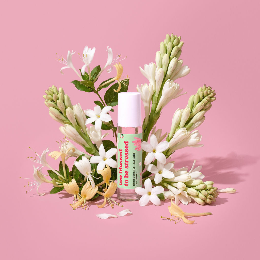 A roll-on bottle labeled 'too blessed to be stressed' surrounded by jasmine flowers on a solid pink background.