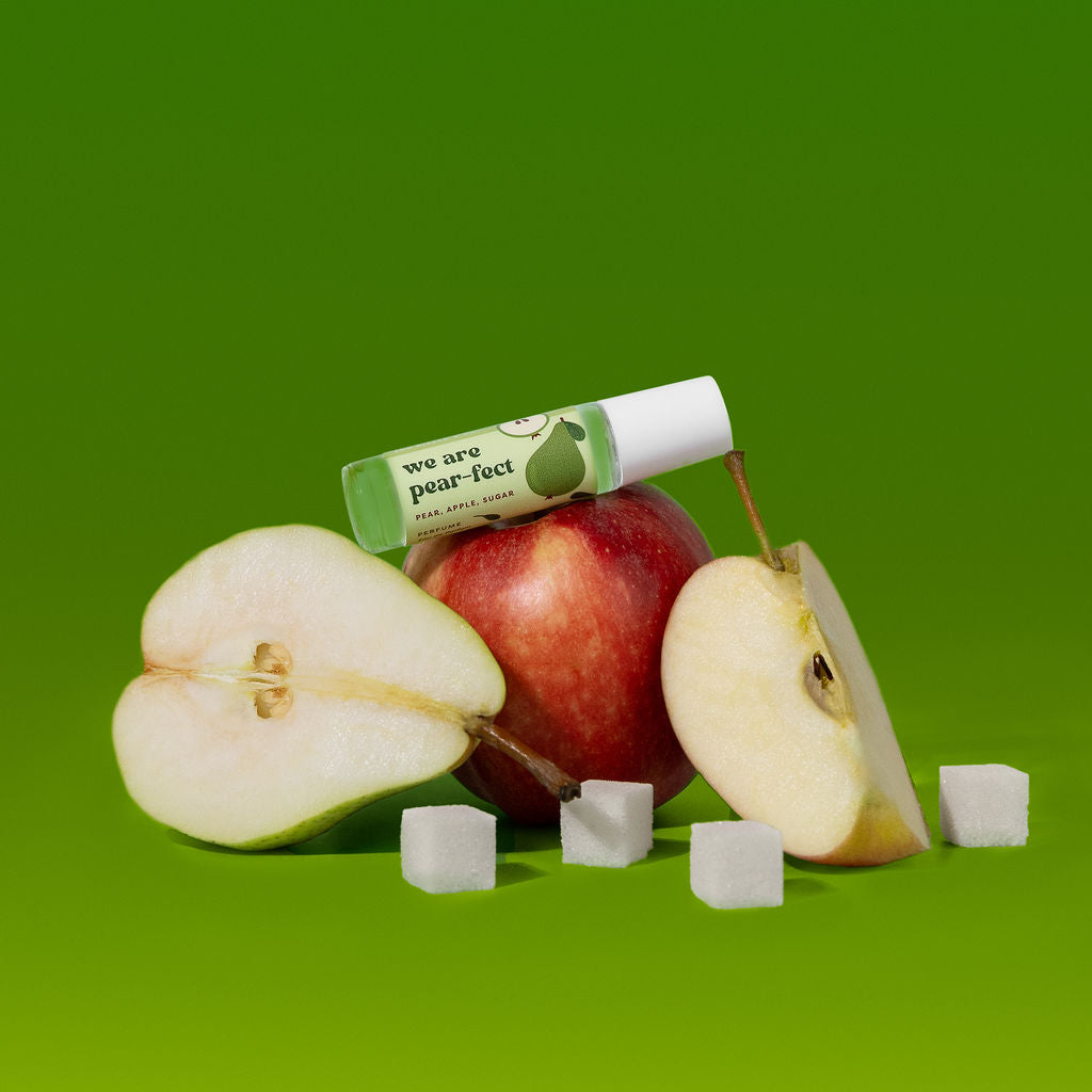 A small clear roll-on bottle labeled 'we are pear-lect' rests on a red apple, surrounded by sliced pears, a whole apple, and sugar cubes against a solid green background.
