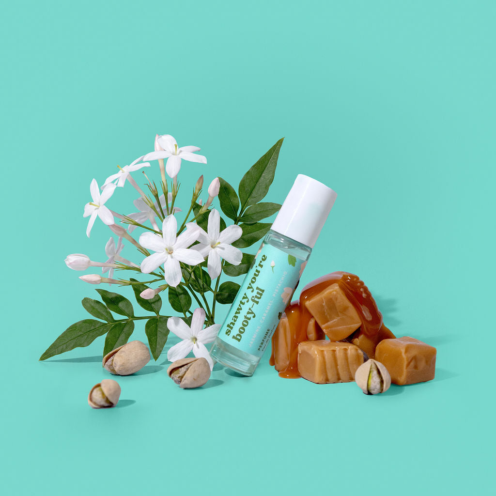 A clear roll-on bottle labeled 'shawty you're booty-ful' with a white cap, styled with white jasmine flowers, green leaves, caramel pieces, and pistachio nuts on a turquoise background.