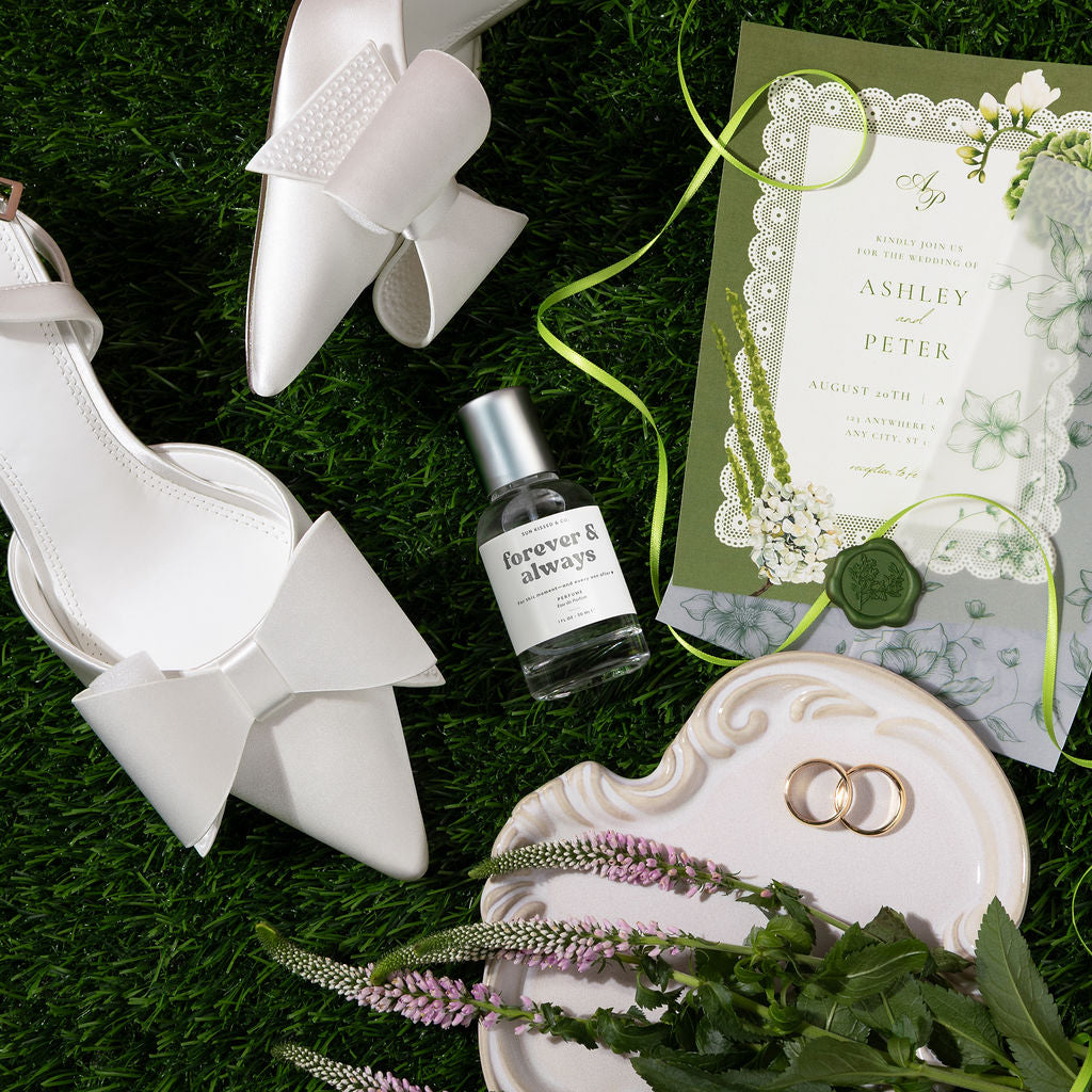 A styled flat-lay featuring a pair of white pointed-toe high heels with large bow details, a small bottle of Forever & Always perfume, a green and white wedding invitation card, two gold rings on a white dish, and green leaves and purple flowers on a textured green grass-like surface.