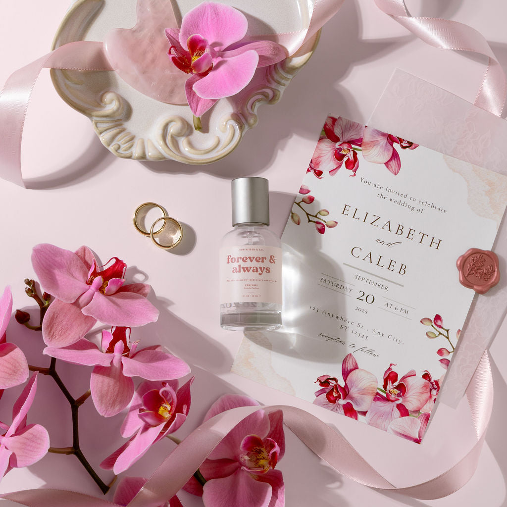 A styled flat-lay featuring a small bottle of 'forever & always' clear perfume with a silver cap, a wedding invitation card with pink orchid illustrations and a wax seal, two gold rings, and pink orchid flowers on a light pink background with a decorative plate and ribbon.