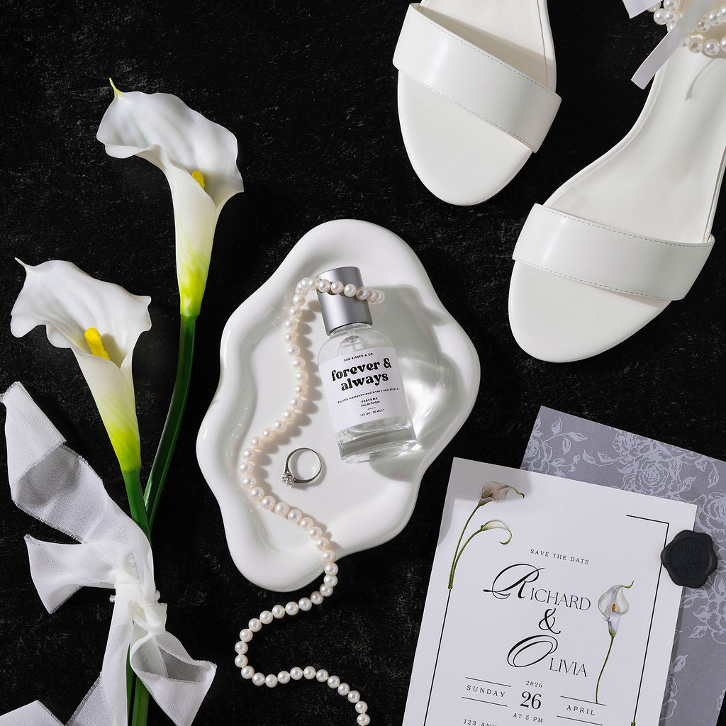 A styled flat-lay featuring a clear glass perfume bottle labeled 'forever & always' with a silver cap, draped with a white pearl strand and a pearl ring on a white ceramic dish. The scene includes white calla lilies, white leather sandals, a wedding invitation card, and a black textured background.