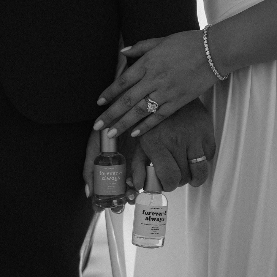 A black and white close-up of a bride and groom holding hands, with the bride's hand resting on the groom's arm. Both are holding Forever 21 'forever & always' perfume bottles.