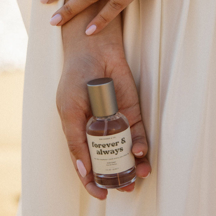 A person in a cream dress holds a small bottle of Forever & Always Eau de Parfum in their hand, with a large silver ring on their finger, against a warm, softly blurred background.